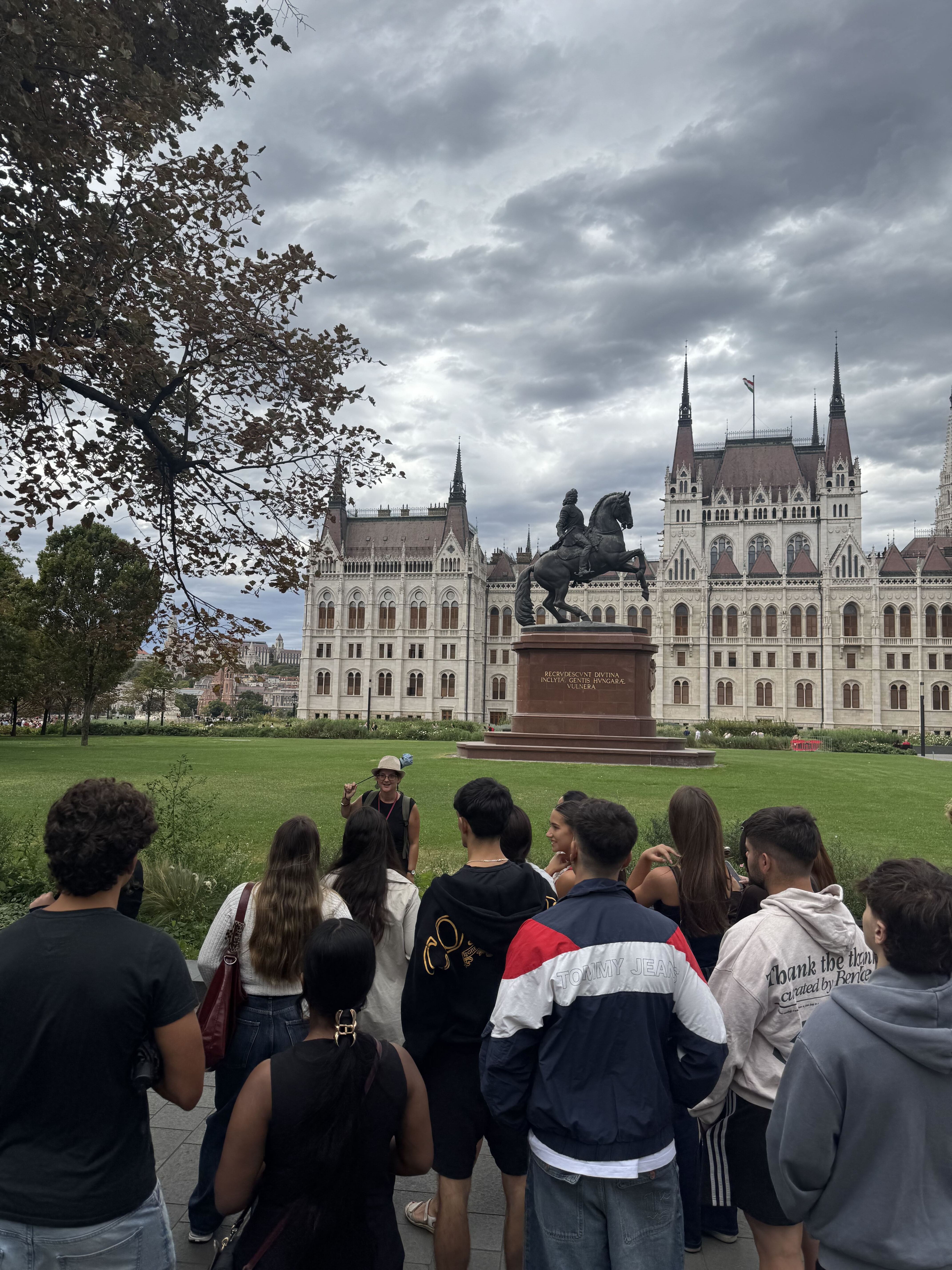 Group of Fuse tenants gathered on the lawn in front of Budapest's Parliament building, listening to their guide, with a bronze equestrian statue and dramatic cloudy sky behind them